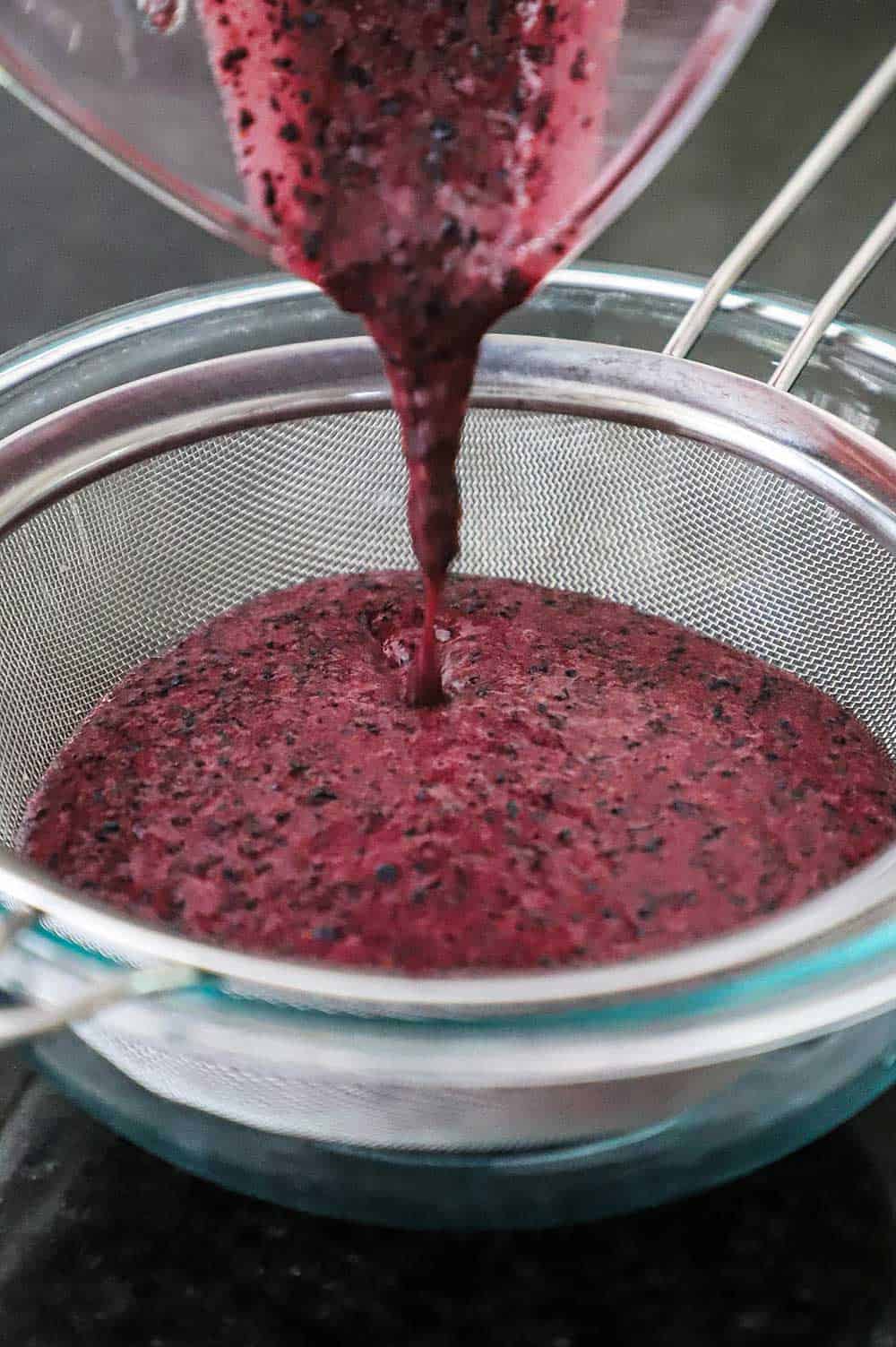 Puréed blueberries being poured into a fine-mesh sieve over a glass bowl.