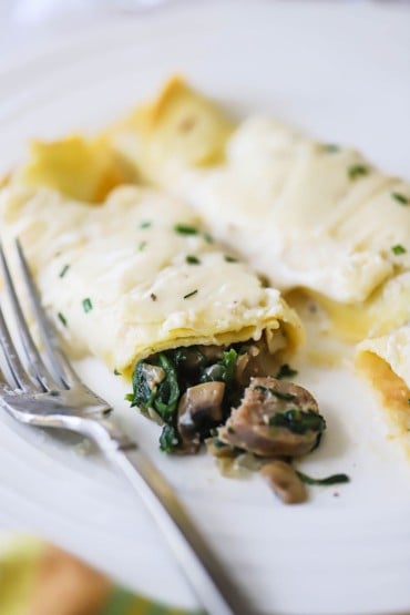 Two savory crêpes on a plate with one that has been broken open with mushrooms and spinach spilling out of it.