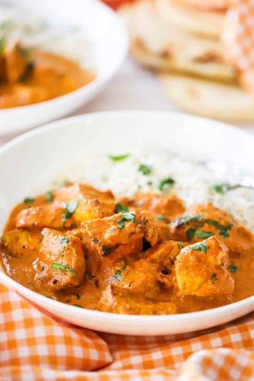 A white bowl filled with chicken tikka masala and steamed white rice with a orange checkered napkin next to it.