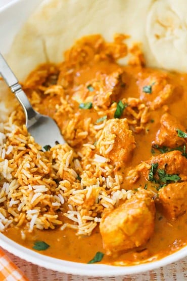 A close-up view of a white bowl filled with chicken tikka masala that has been combined with white rice with a fork.