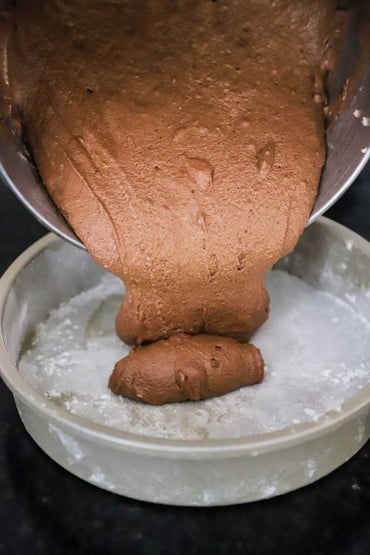 Chocolate cake batter being poured from a mixing bowl into a greased 9-inch cake pan.