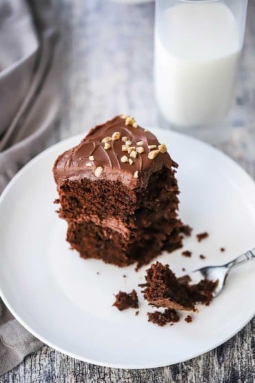 A slice of chocolate cake that has been half eaten sitting on white plate next to a glass of milk.