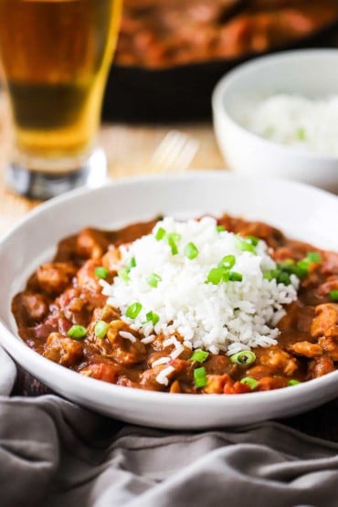 A close-up view of a white bowl filled with creole chicken with steamed white rice on top garnished with chopped scallions.