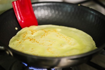 A homemade crêpe in a small non-stick skillet with a red spatula about to flip it.