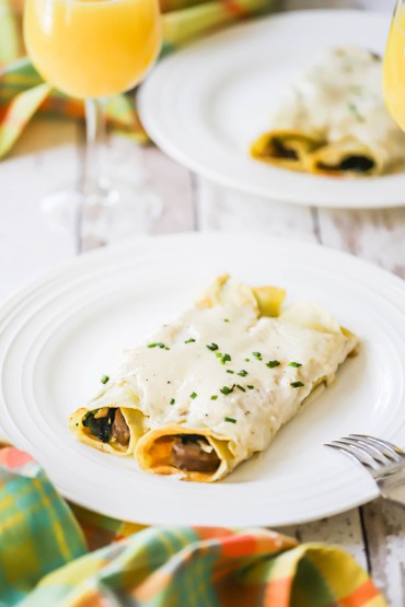 A white dinner plate filled with two untouched savory crêpes topped with a creamy mornay sauce and snipped chives.