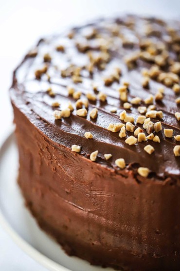 A close-up view of a classic chocolate cake sitting on a white circular platter and is topped with toffee bits.