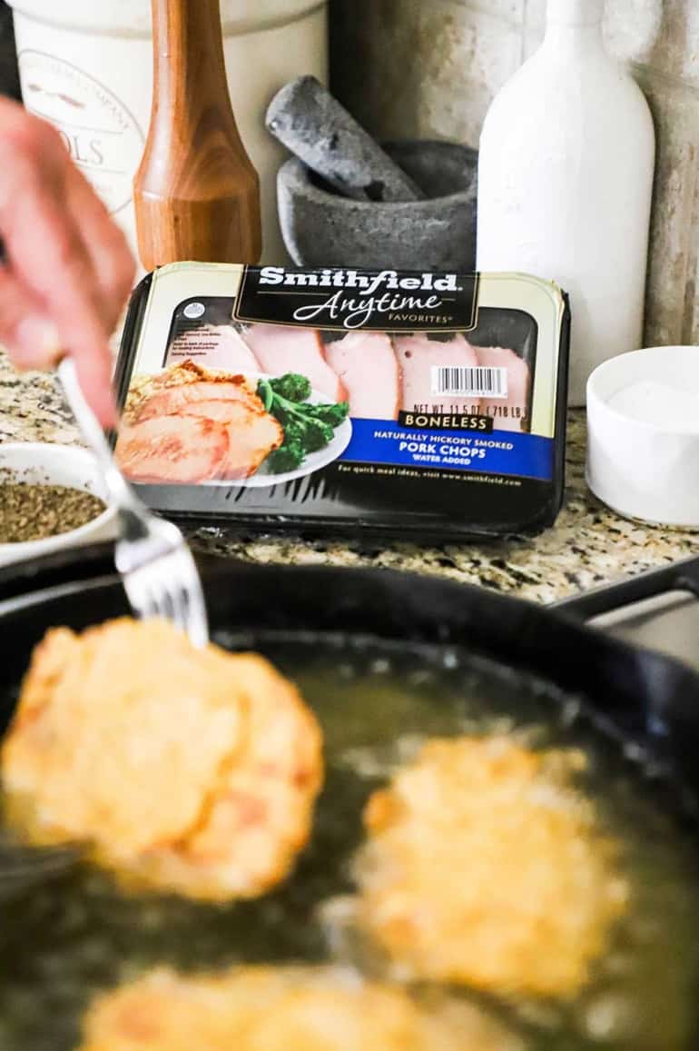 A cast-iron skillet in the foreground filled with hot oil and fried pork chops sizzling and a package of Smithfield pork chops in the background.