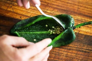 A person using a small spoon to scrap out the seeds from inside a roasted poblano pepper that has been split open.