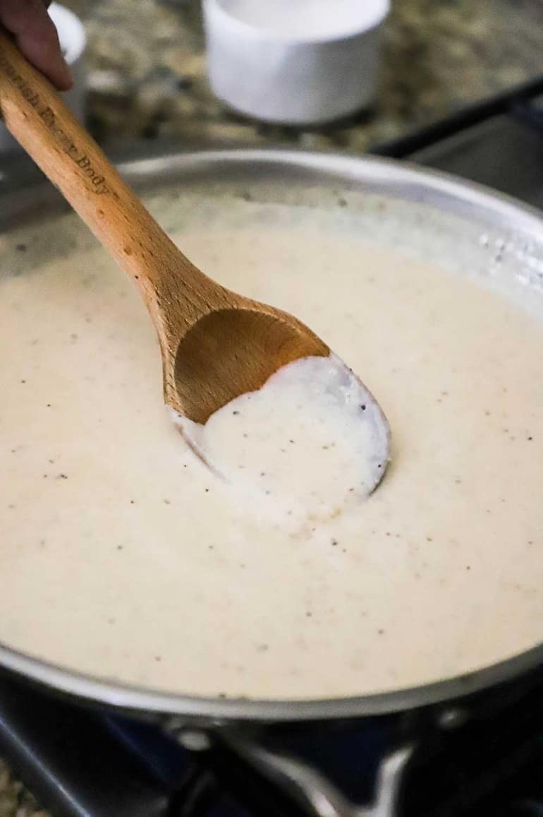 A large silver skillet filled with cream gravy with a wooden spoon inserted into the middle of it.