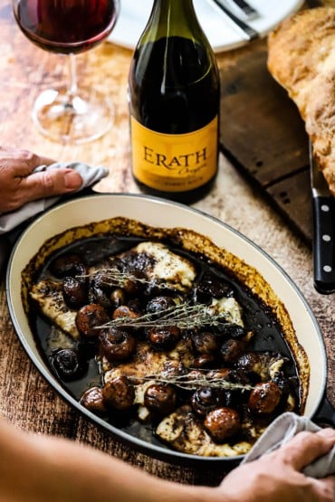 A person holding onto the sides of an oval baking dish that is filled with seared haddock with agrodolce sauce with a bottle of Erath wine nearby.