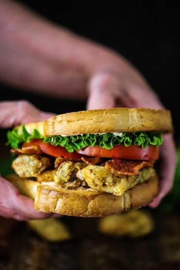 A person holding a Fried Oyster BLT consisting of two toasted slices of bread with crunchy oysters, crispy bacon, sliced tomatoes, and lettuce.