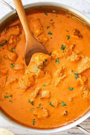 An overhead view of a large silver saucepan filled with creamy chicken tikka masala with a wooden spoon resting in the sauce.