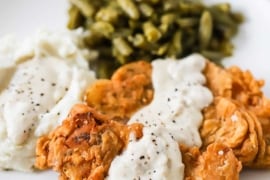 A close-up view of two Southern-fried pork chops on a dinner plate with cream gravy drizzled over the top of them and sitting next to mashed potatoes and green beans.