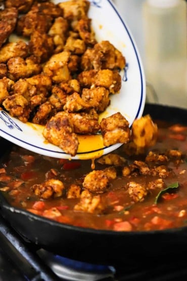 Fried chunks of seasoned chicken being transferred into a cast-iron skillet filled with a red sauce and chopped vegetables.