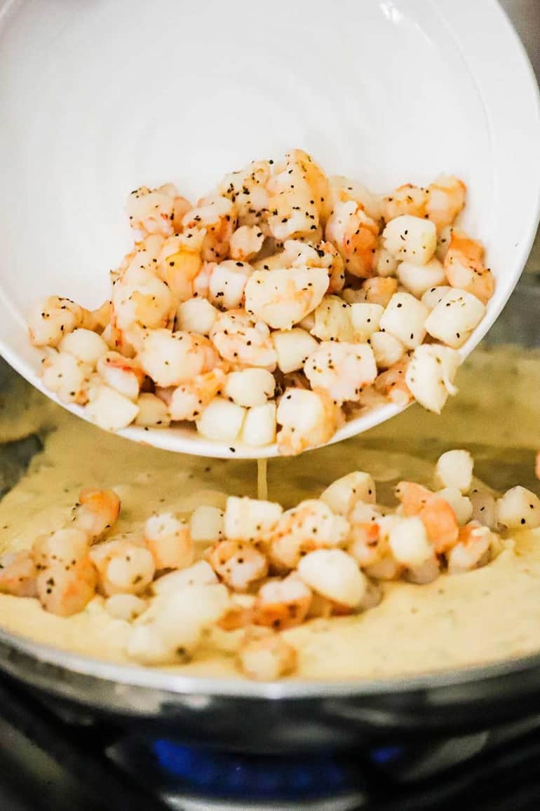 Cook pieces of shrimp and scallops being transferred into a skillet filled with a cream sauce.