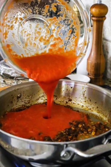 Puréed tomatoes being poured from a glass bowl into a skillet with sautéd vegetables.