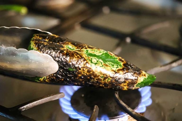 A pair of metal tongs holding a poblano pepper over a gas flame on a stovetop.