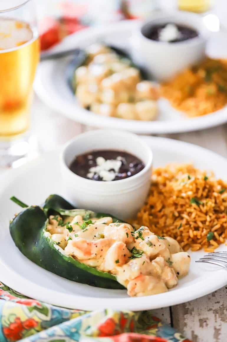 Two plates holding seafood stuffed poblano peppers sitting next to rice and beans and a glass of beer nearby.