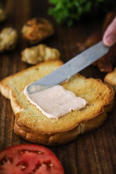 A person using a butter knife to spread sriracha mayonnaise onto a slice of toasted white bread.