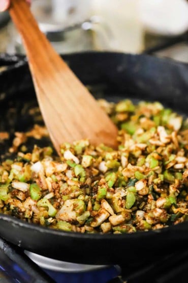 A wooden spatula being used to stir chopped onions and peppers as they sauté in a black skillet.