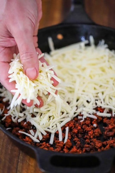 A person sprinkling shredded Monterey Jack cheese into a cast-iron skillet filled with cooked crumbled chorizo sausage.