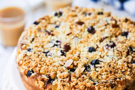 A straight-on view of a circular blueberry almond coffee cake sitting on a raised white cake stand.