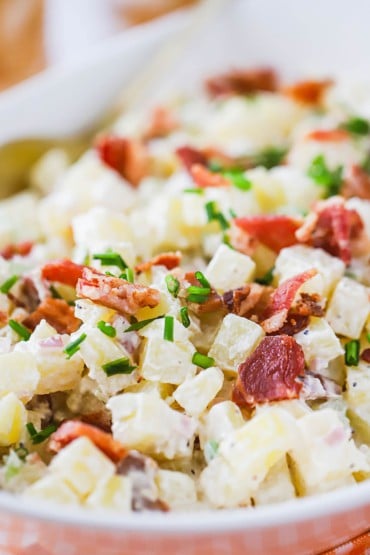 A close-up view of a white bowl filled with creamy horseradish potato salad topped with bacon pieces and snipped chives.