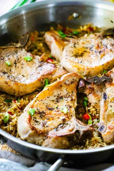 A straight-on view of a large stainless steel skillet filled with pan-seared pork chops on top of seasoned rice.