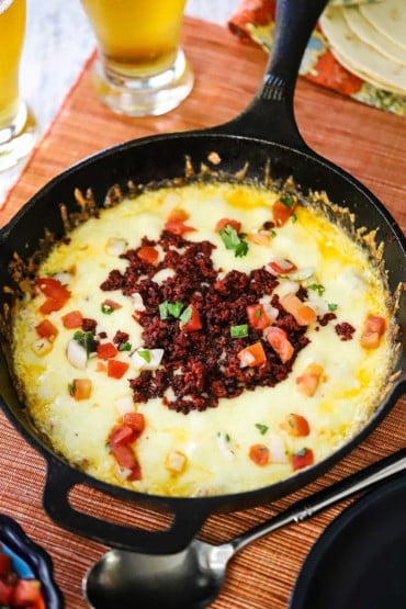 A black cast-iron skillet filled with melted white cheese and topped with cooked chorizo and chopped tomatoes.
