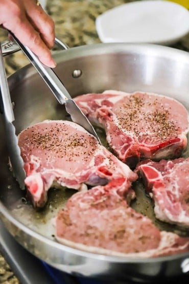 A person using a pair of metal tongs to place uncooked bone-in pork chops into a large stainless steel skillet.