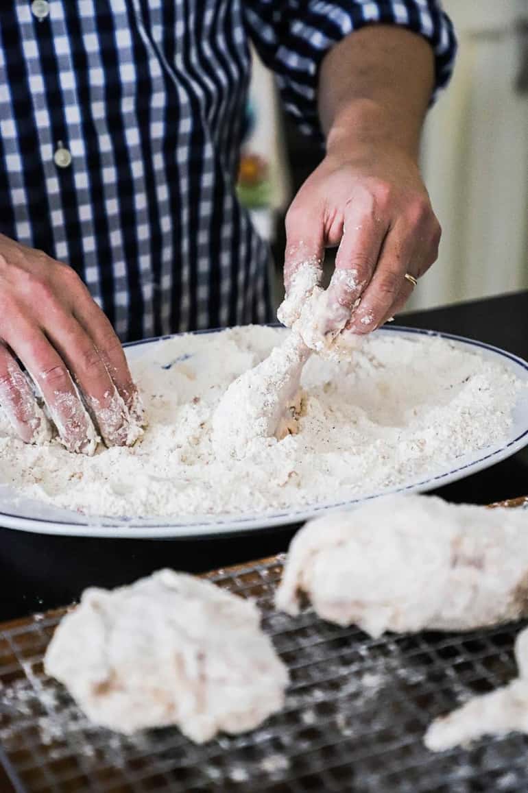 A person using his hands to dredge a chicken leg in seasoned flour on a white platter.