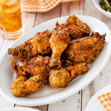 A large oval white platter filled with a stack of Southern-fried chicken sitting next to a glass of iced tea and a checkered napkin.