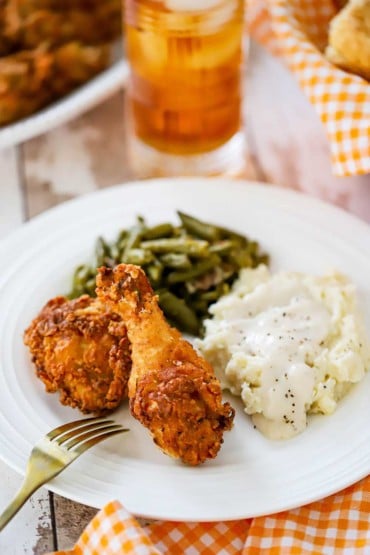 A white dinner plate filled with pieces of Southern fried chicken, a pile of mashed potatoes with cream gravy, and a helping of green beans.