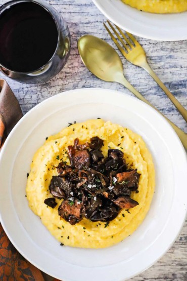 An overhead view of a white bowl filled with creamy polenta and caramelized onions and wild mushrooms next to a glass of red wine.