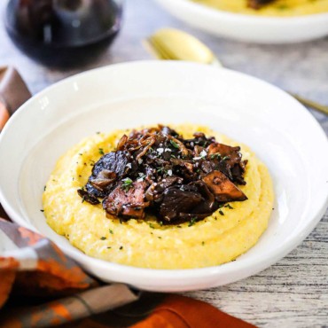 A close-up view of a white serving bowl filled with a mound of creamy polenta and topped with caramelized onions and porcini mushrooms.