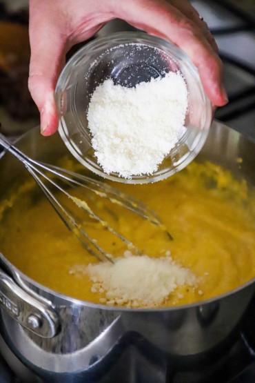 A person dumping grated Parmesan cheese into a silver saucepan filled with creamy polenta.