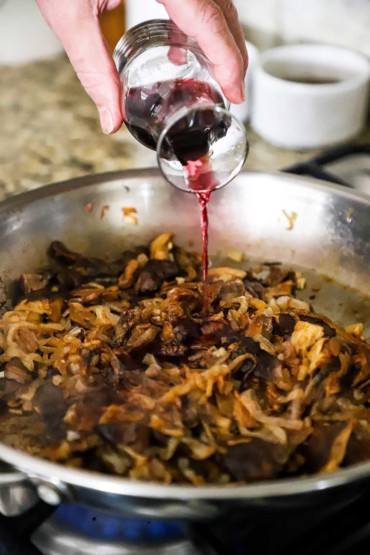 A person pouring red wine from a small wine carafe into a skillet filled with caramelized onions and sautéed mushrooms.