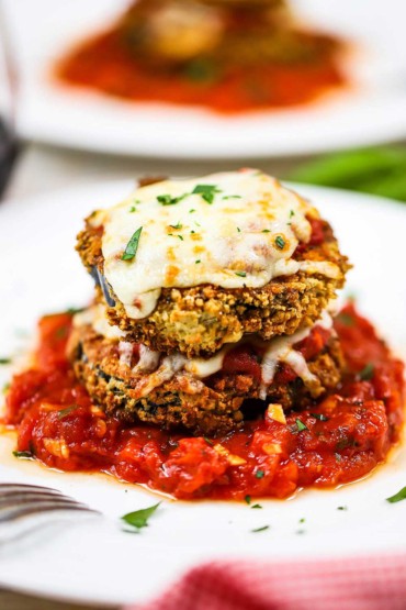 A close-up view of two eggplant medallions that have been breaded and air-fried then baked with cheese and then stacked on a bed of marinara on a white dinner plate. 