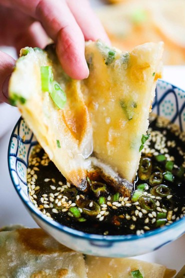 A person dipping a quarter of a scallion pancake into a patterned bowl filled with a soy dipping sauce.