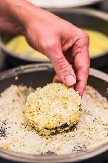 A person holding up an eggplant medallion that has been dredged through flour, then egg, then a pan of breadcrumbs.