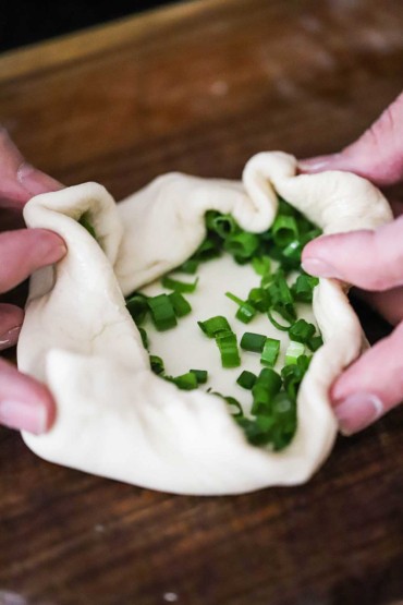 A person folding the edges of a disc of dough with sliced scallions in the middle.