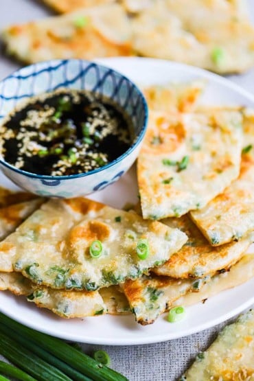 Scallion pancakes that have been quartered and placed on a plate with a bowl filled with a dipping sauce off to the side.