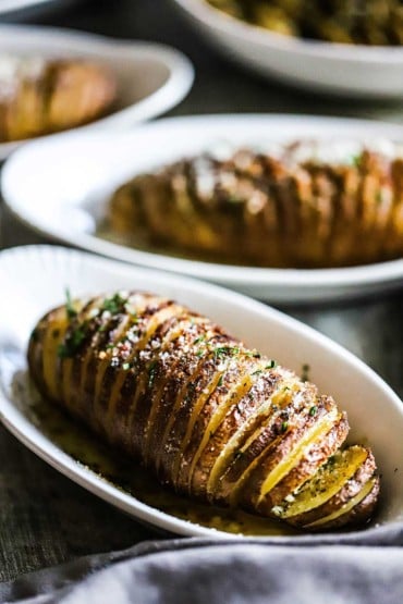 A close-up view of a potato that is prepared in the Hasselback style with thin slices cut into an sprinkled with cheese and herbs.