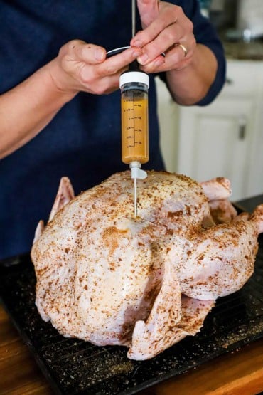 A person using a large meat syringe to inject a butter marinade into a seasoned raw turkey. 
