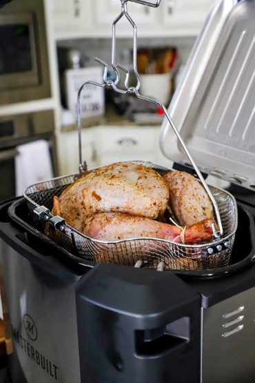 An uncooked seasoned turkey sitting in a wire basket and slowly being lowered into an indoor electric deep-fryer. 