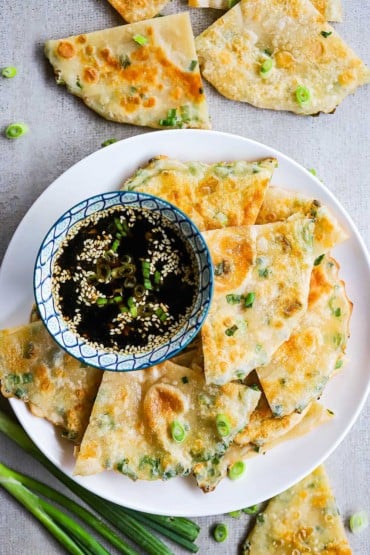 An overhead view of quarted scallion pancakes on a plate with a bowl filled with a dipping sauce.