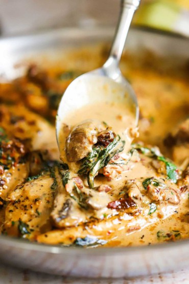 A spoon being used to ladle creamy sauce with spinach and sun-dried tomatoes over seared chicken in a skillet.