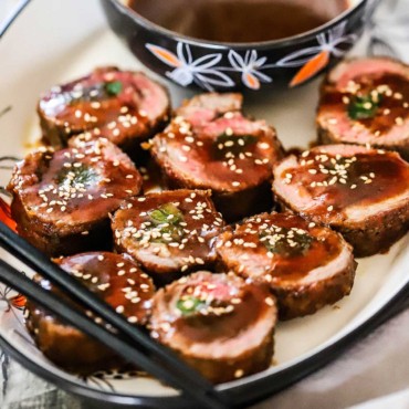 A close-up view of slices of beef negimaki on a platter topped with a brown sauce and sesame seeds.