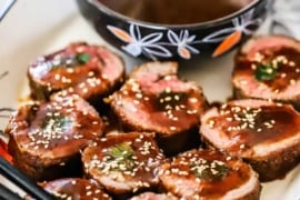 An oval platter filled with pieces of beef negimaki sitting on their sides topped with brown sauce and sesame seeds all sitting next to a bowl of the sauce.