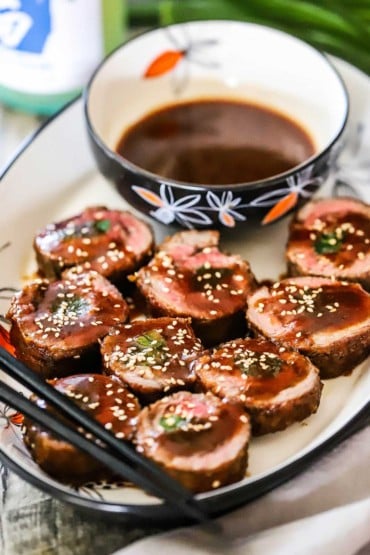 An oval platter filled with pieces of beef negimaki sitting on their sides topped with brown sauce and sesame seeds all sitting next to a bowl of the sauce.